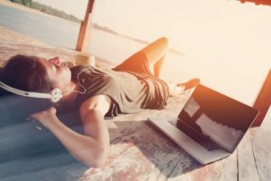 Young woman chilling with laptop and listening music with headphones in shadow shelter. Blurry effect, lens flares effect, intentional sun glare